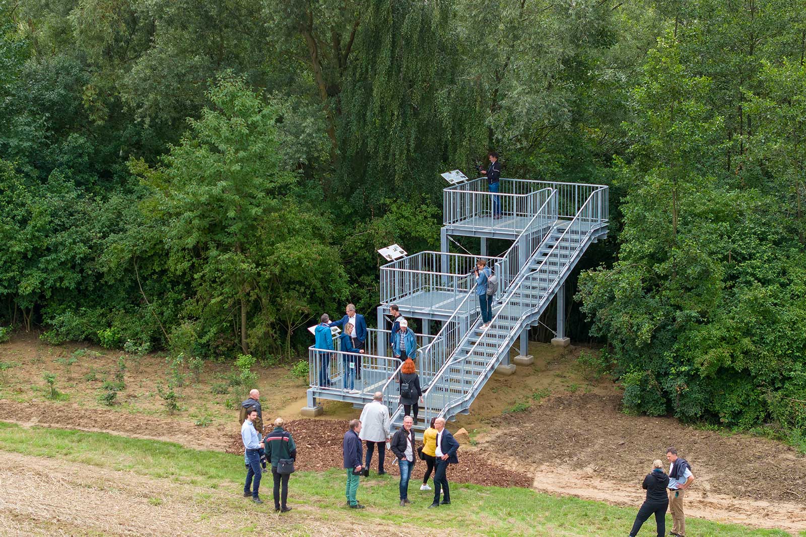 Eine Gruppe von Menschen steht um eine Treppe aus Metall, die in einem grünen Wald steht. Einige lesen Informationen auf Tafeln.