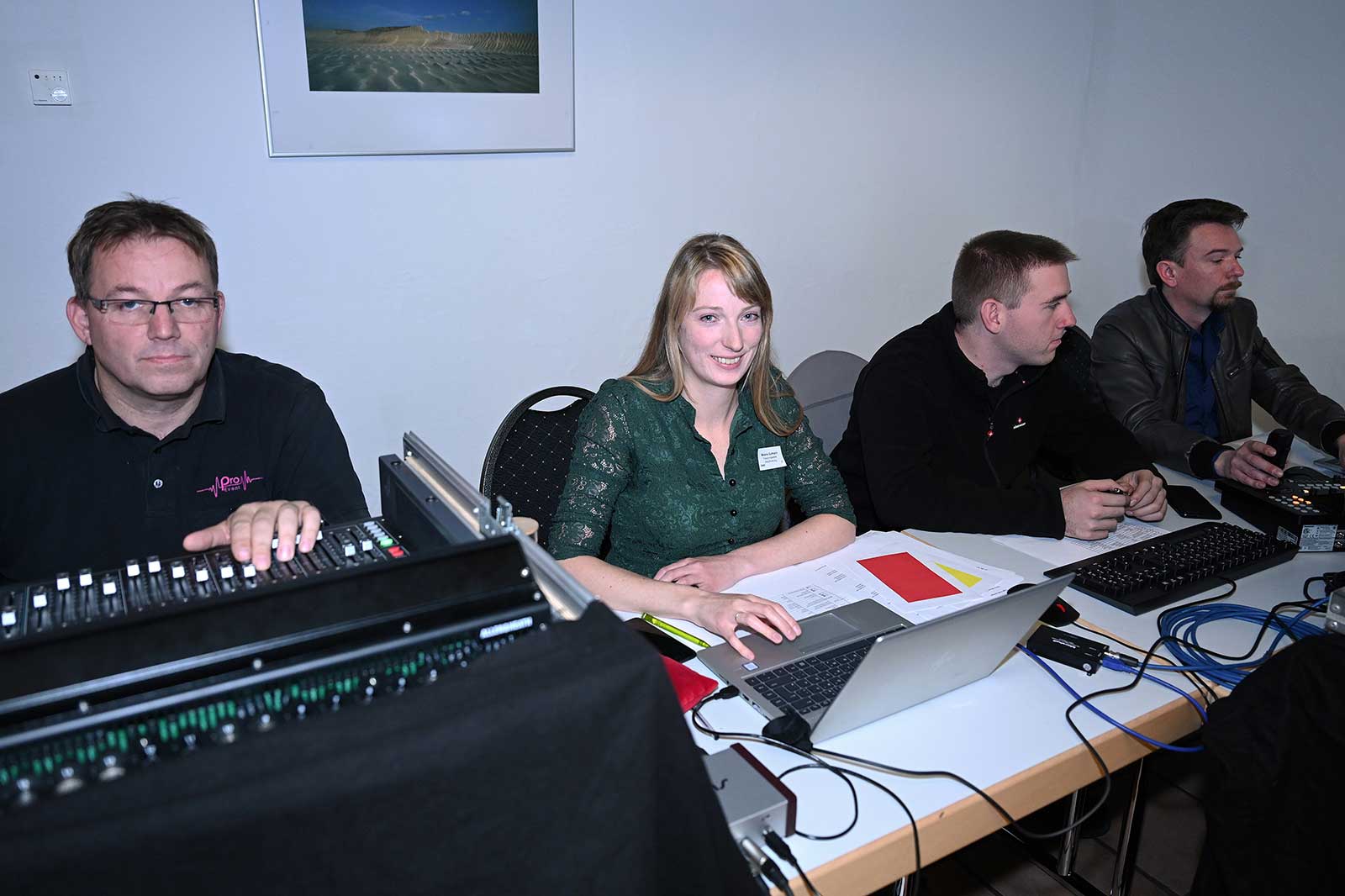 Ein Team arbeitet an Computern und Geräten in einem Raum, konzentriert auf Aufgaben. Papiere und Kabel sind auf dem Tisch sichtbar.