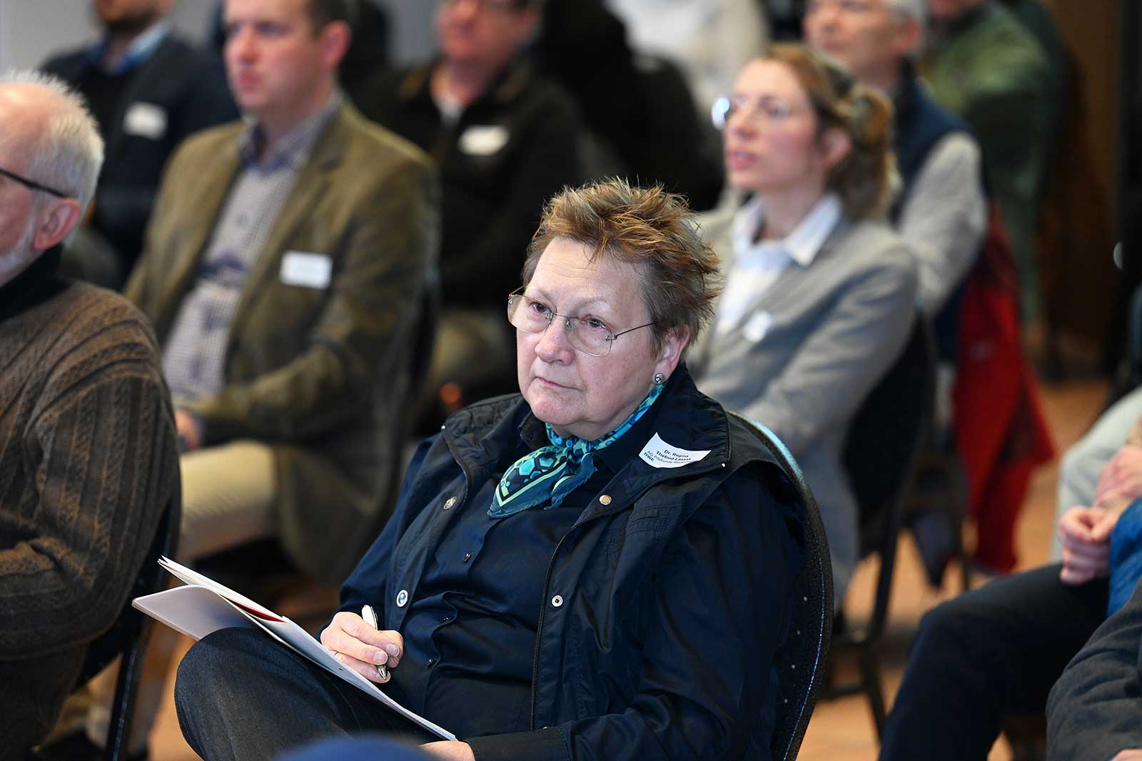 Ein sitzendes Mitglied des Publikums, das eine schwarze Weste und einen blauen Schal trägt und während einer Diskussion Notizen macht.