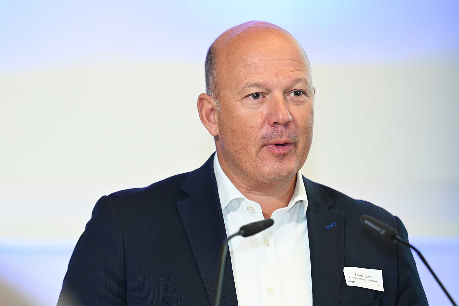 Ein Mann in einem dunklen Blazer spricht an einem Podium mit einem Mikrofon. Ein Namensschild lautet 'Frank Rack, Limited Urban City Vision'.