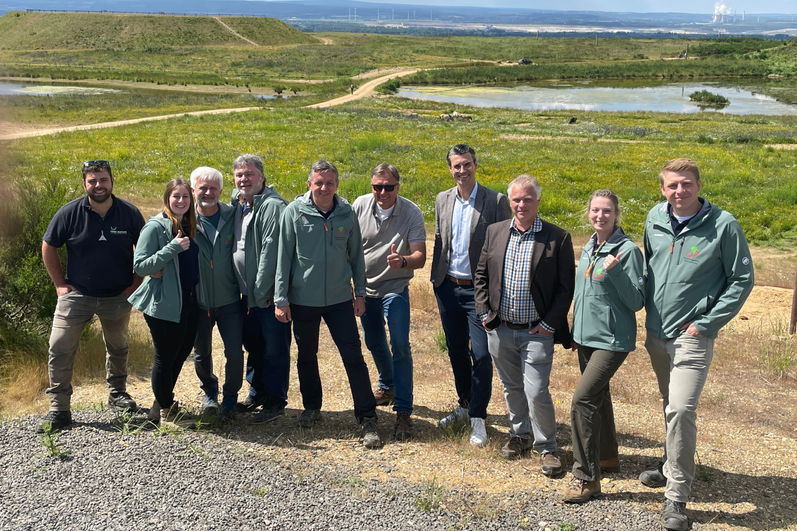 Eine Gruppe von Menschen in grünen Jacken, die im Freien mit einer malerischen Landschaft im Hintergrund posiert, die Hügel und ein Gewässer zeigt.
