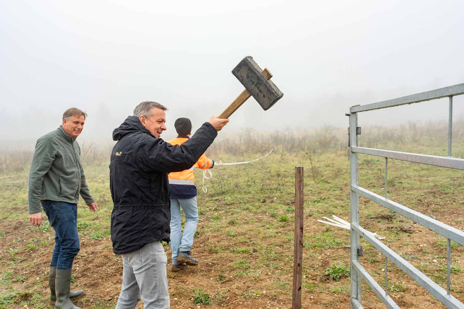 Drei Männer arbeiten im Nebel, einer schlägt mit einem Hammer auf einen Zaunpfosten. Der Boden ist grasig und feucht.