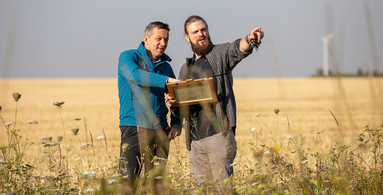 Zwei Männer stehen in einem Feld und zeigen auf ein Objekt, während sie einen Holzrahmen mit Messungen präsentieren. Windräder sind sichtbar.