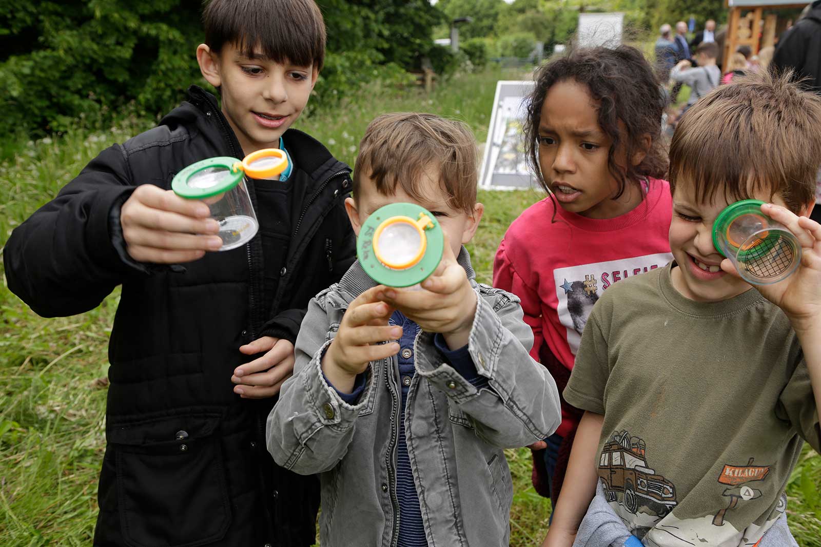 Vier Kinder untersuchen etwas draußen in der Natur mit grünen und orangen Lupenbehältern.