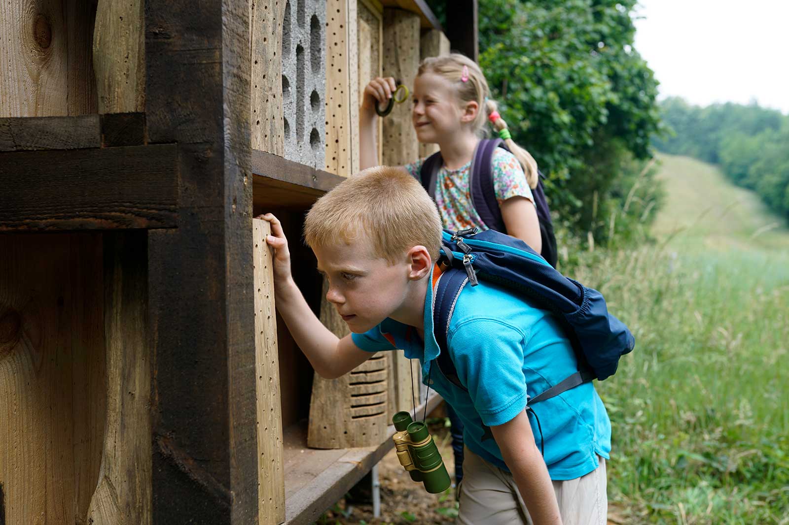Zwei Kinder schauen neugierig in eine Holzstruktur in einer natürlichen Umgebung, einer hält ein Fernglas.