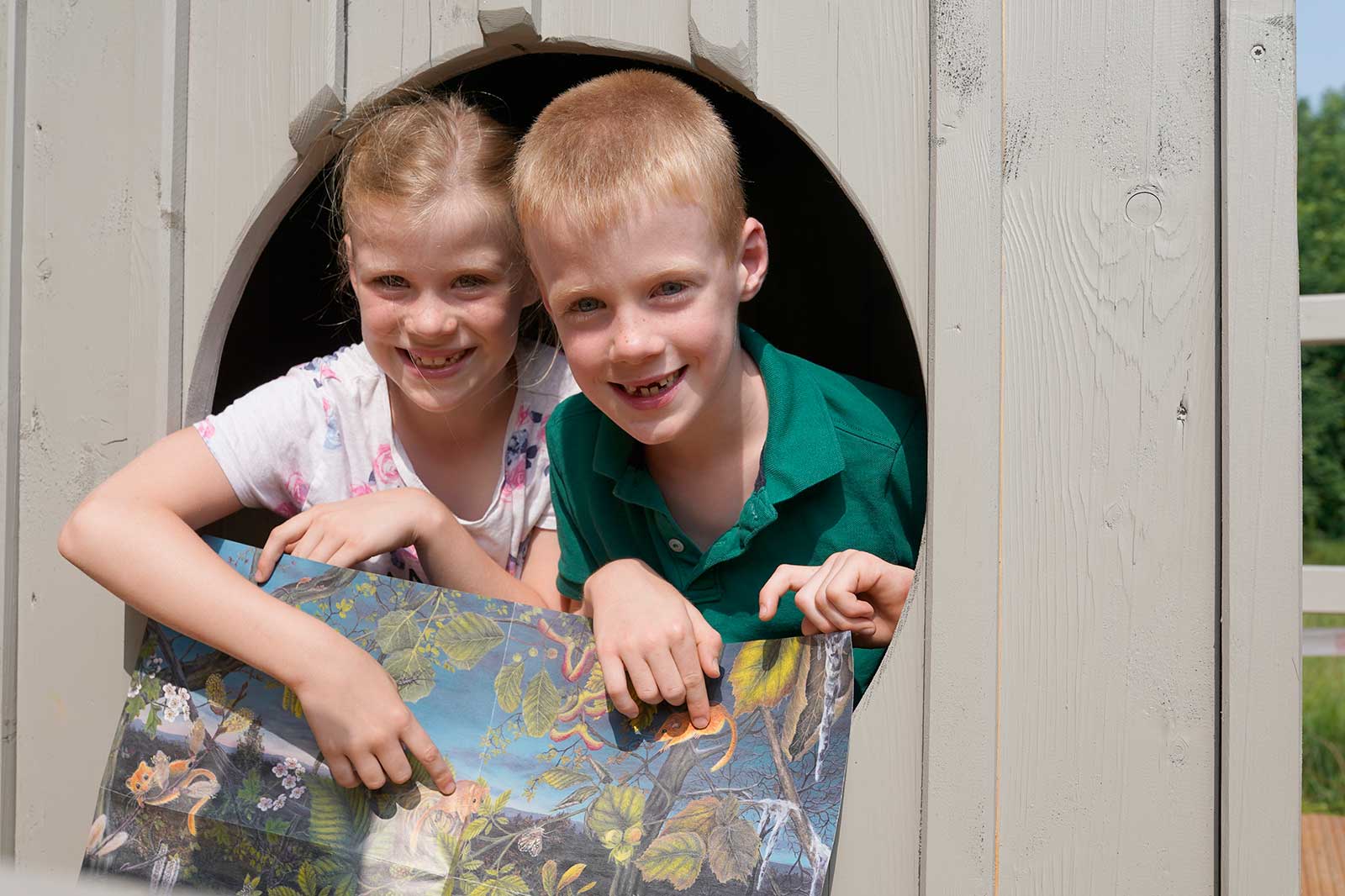 Zwei Kinder schauen durch eine runde Öffnung in einem Holzspielhaus und halten ein buntes illustriertes Poster von Tieren.