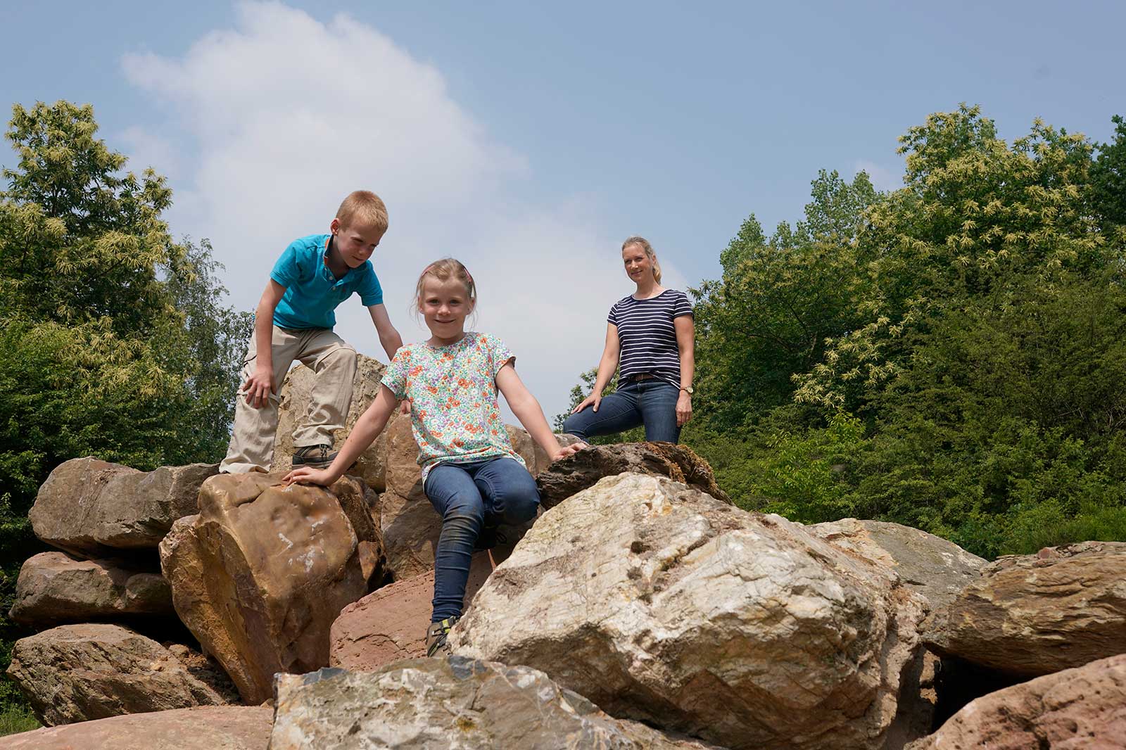 Drei Kinder klettern auf großen Steinen in einem grünen Bereich unter einem blauen Himmel und genießen einen sonnigen Tag im Freien.