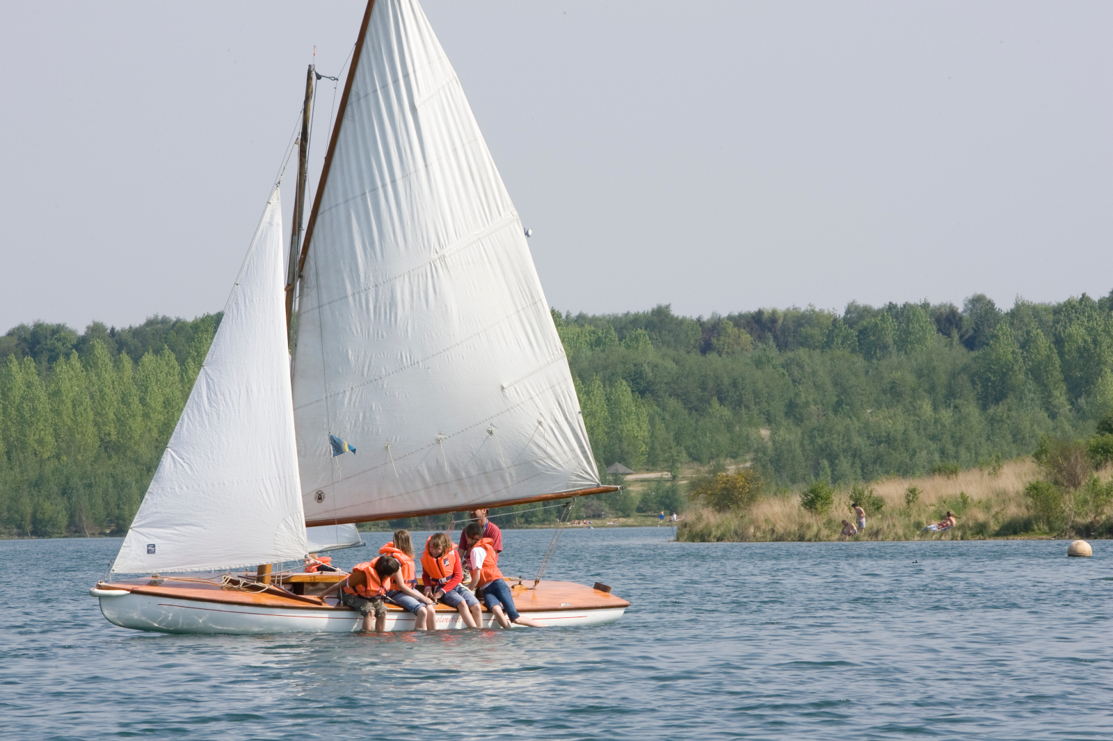 Ein Segelboot mit vier Personen in orangefarbenen Schwimmwesten auf einem ruhigen See, umgeben von grünen Bäumen im Hintergrund.