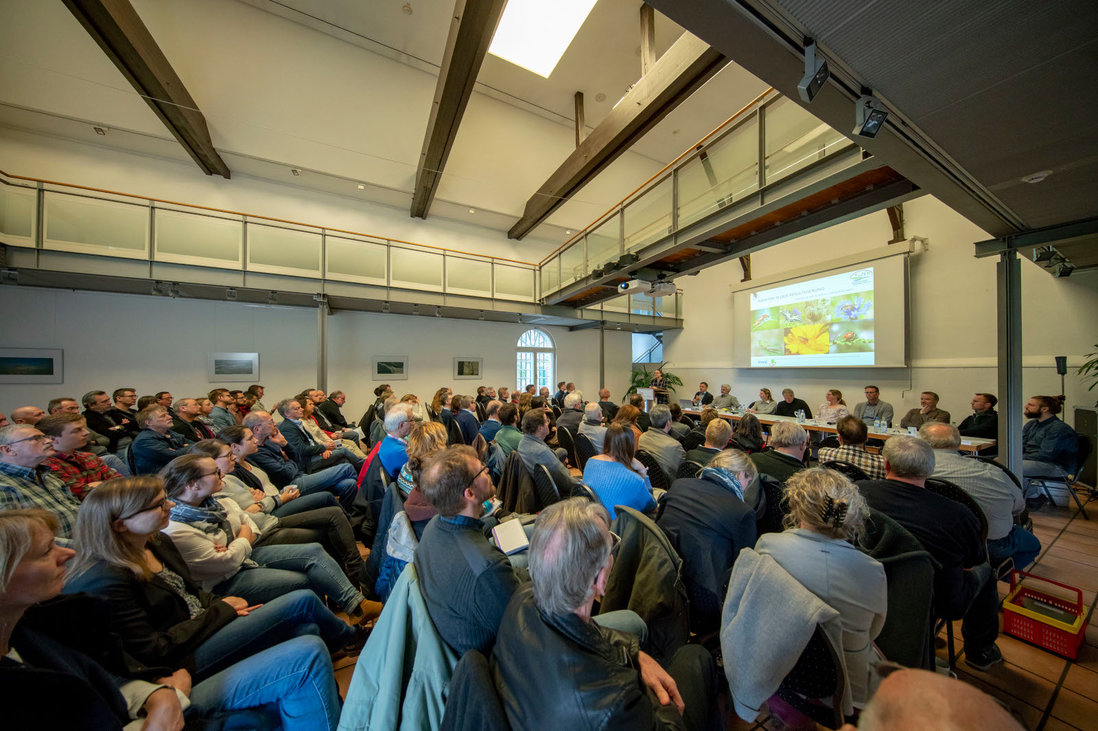 Ein großes Publikum hört aufmerksam einer Podiumsdiskussion in einem geräumigen Raum mit Holzstrahlen und einer Präsentationsleinwand zu.