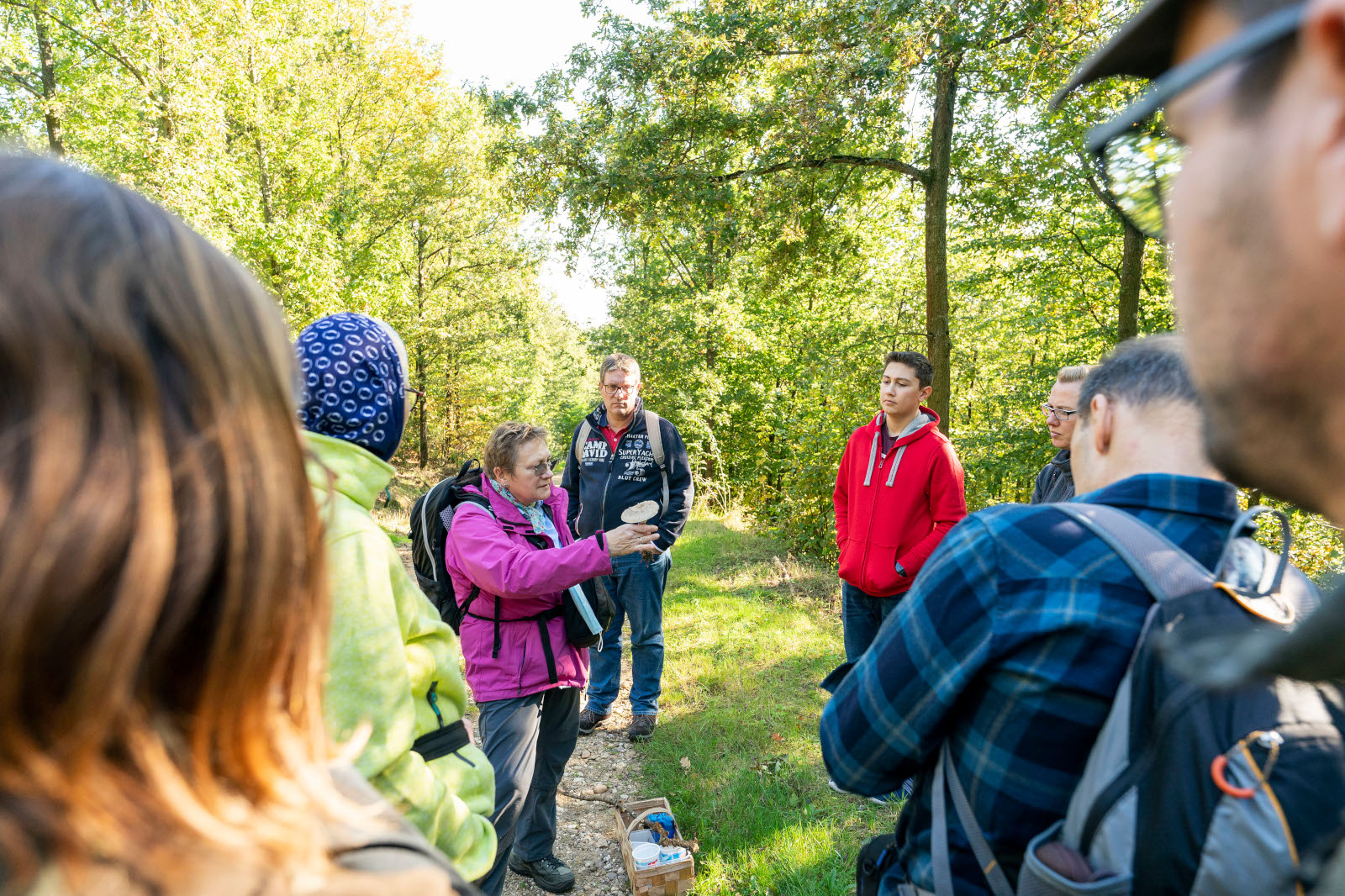 Eine Gruppe von Menschen in einem Wald hört einem Sprecher in einer pinken Jacke zu, umgeben von Grün und Sonnenlicht.