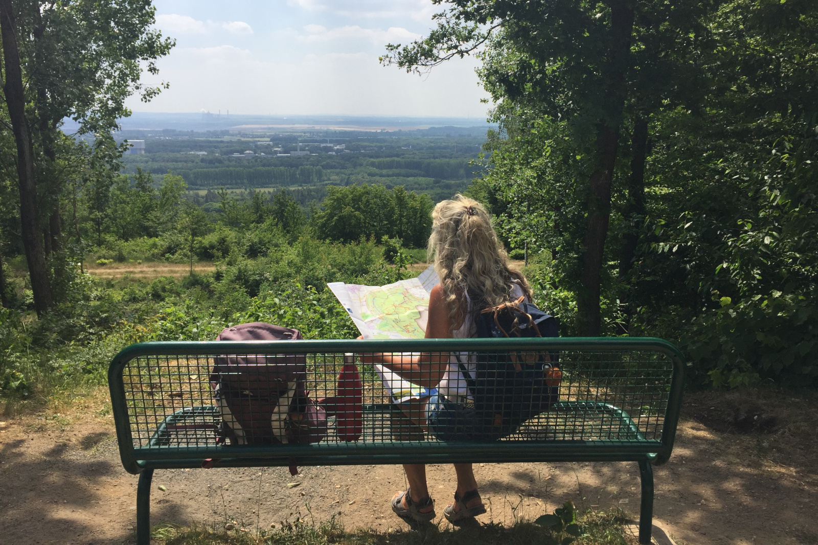 Eine Frau mit langen Haaren sitzt auf einer grünen Bank und studiert eine Karte mit einer malerischen Aussicht auf grüne Bäume und Felder.