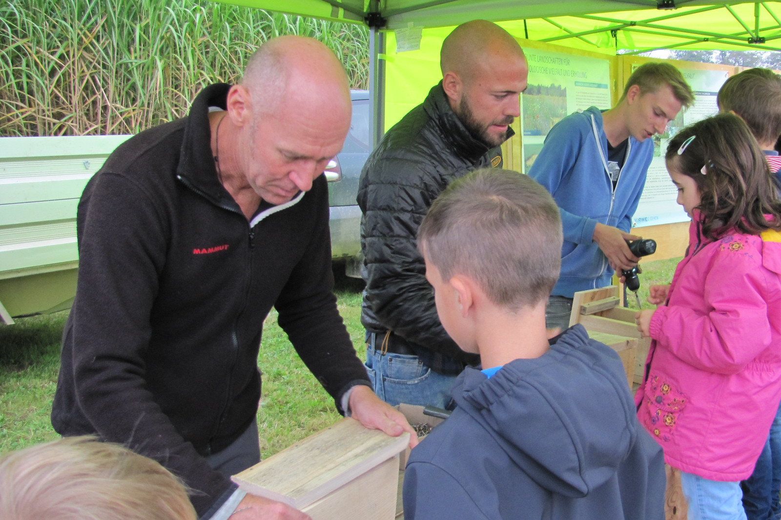 Kinder und Erwachsene beteiligen sich an einer Bildungsaktivität unter einem grünen Zelt. Sie bauen Holzkästen für die Natur.