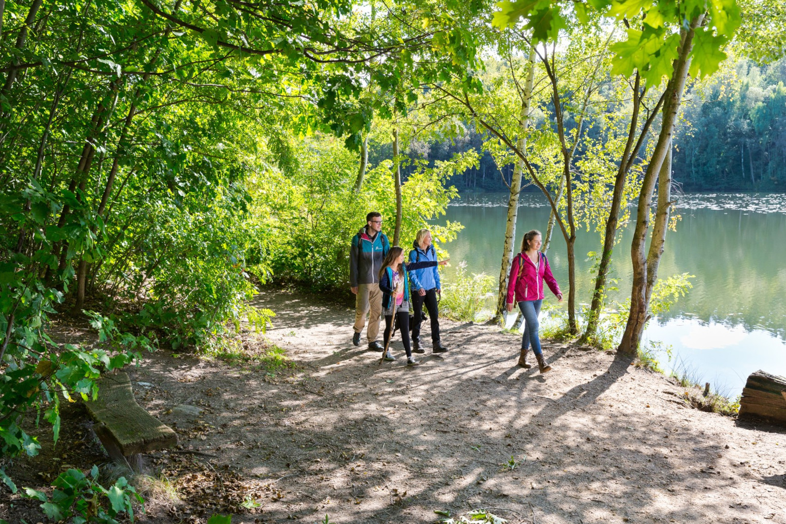 Eine Familie geht einen schattigen Weg entlang eines friedlichen Sees, umgeben von Bäumen und Grün in einer sonnigen Umgebung.