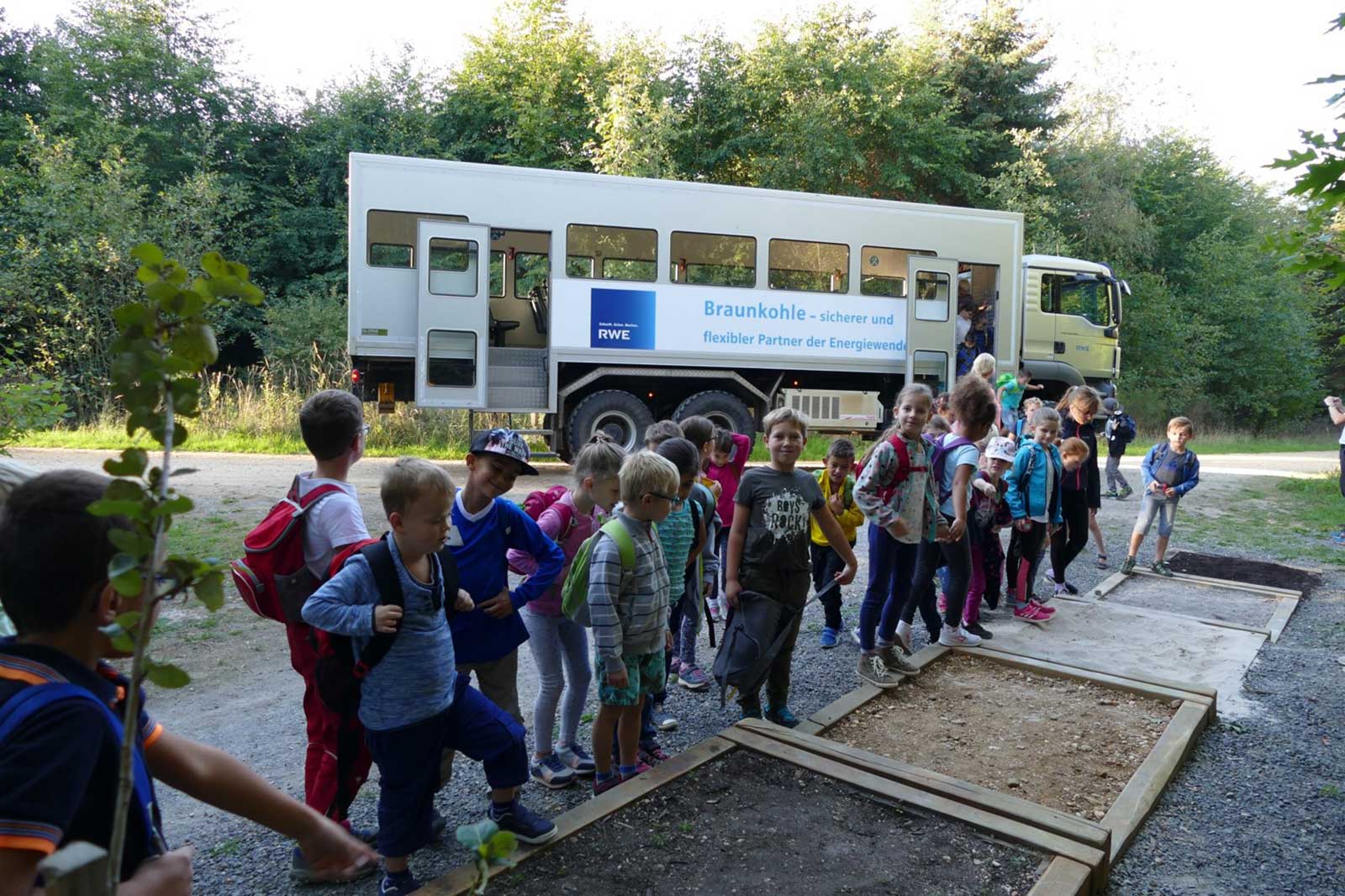 Eine Gruppe von Kindern versammelt sich in der Nähe eines Lastwagens mit dem Logo von RWE und interessiert sich für eine Bildungsaktivität im Freien.