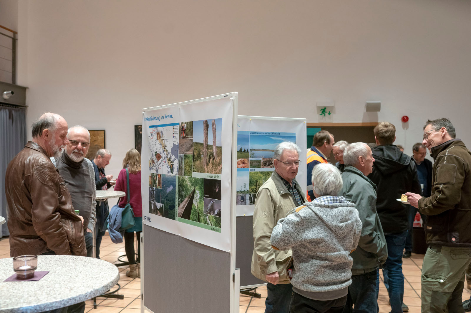 Eine Gruppe von Menschen versammelt sich um Informationsplakate und diskutiert über die Rekultivierung im Revier. Tische mit Getränken sind vorhanden.