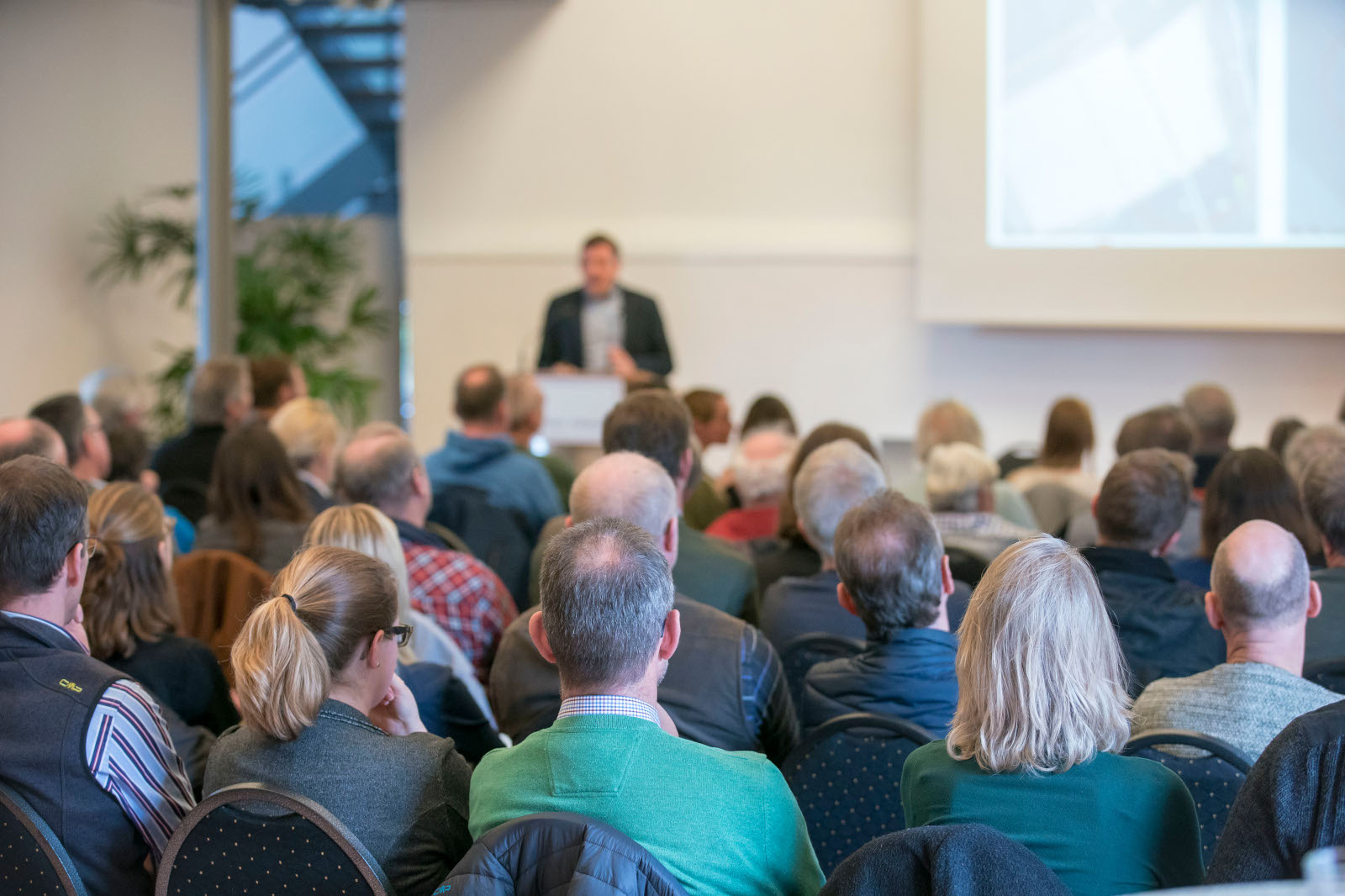 Eine Menschenmenge hört aufmerksam einem Redner in einem gut beleuchteten Raum mit Pflanzen im Hintergrund zu.