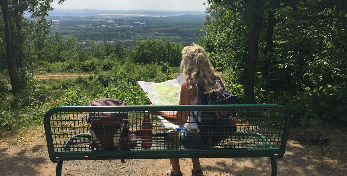 Eine Frau mit lockigem Haar sitzt auf einer Bank, studiert eine Karte und blickt auf eine grüne Landschaft unter einem sonnigen Himmel.