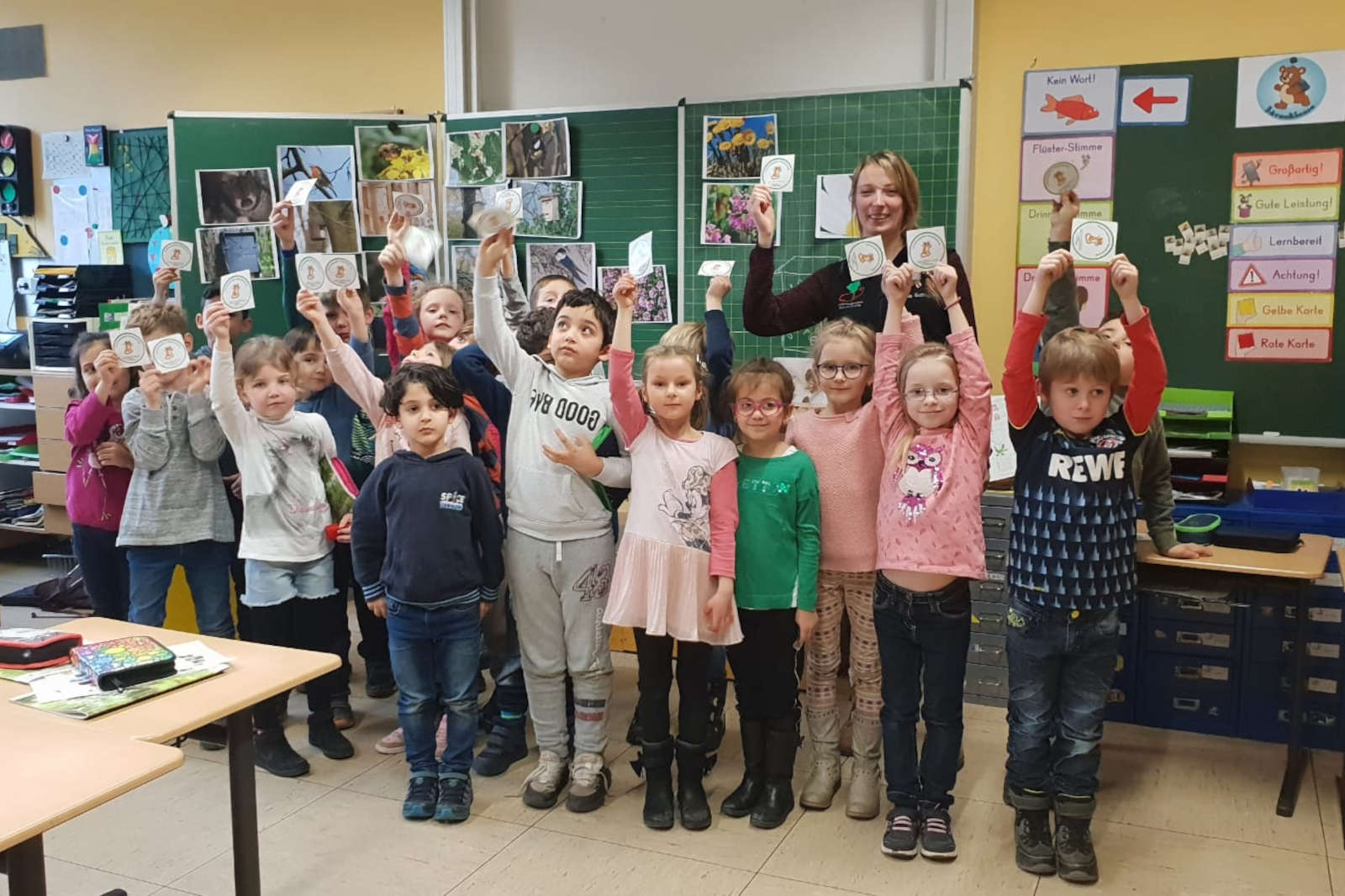 Eine Gruppe von Kindern hält in einem Klassenraum Karten hoch, während Lehrmaterialien an den Wänden sichtbar sind.