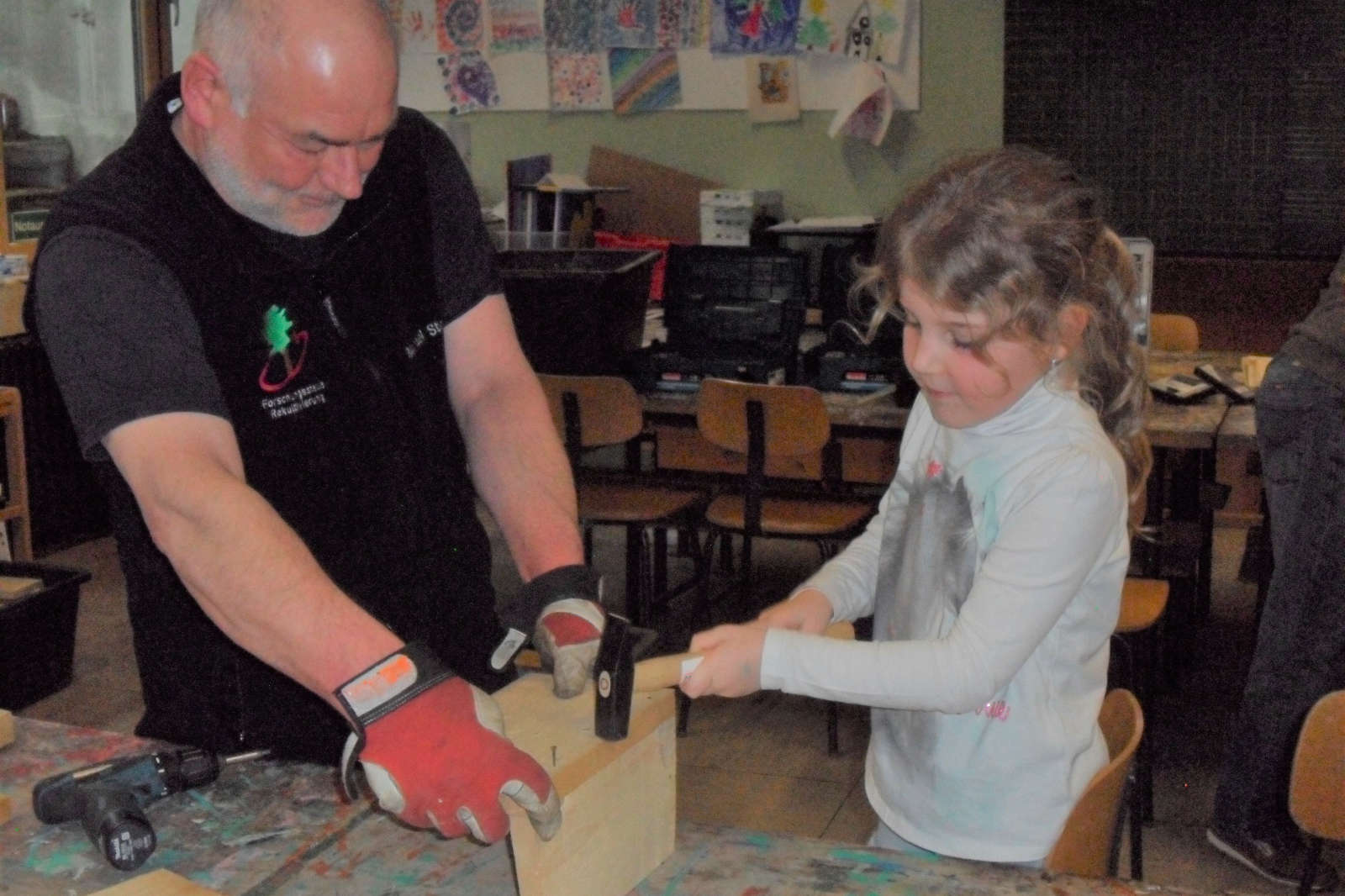 Ein Erwachsener und ein Kind arbeiten in einem Klassenzimmer gemeinsam an einem Holzprojekt, während sie Werkzeuge auf einem Holzblock verwenden.