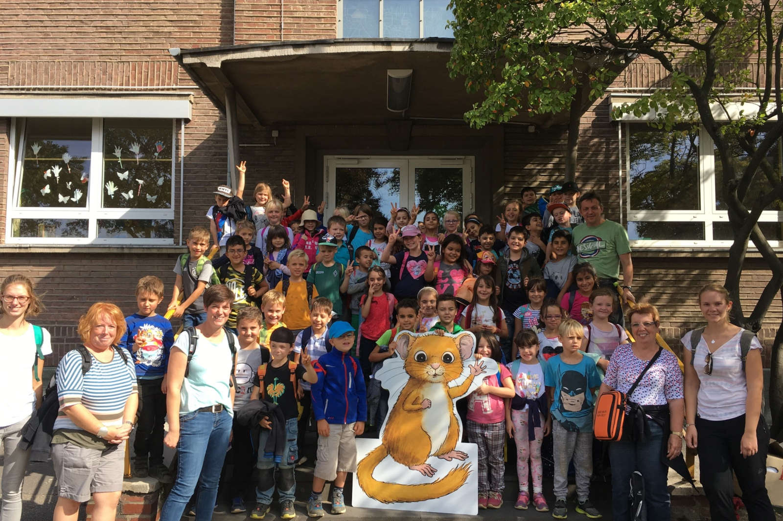 Eine Gruppe von Kindern steht vor einer Schule, mit einem großen Cartoon-Tiercharakter zu ihren Füßen und dekorierten Fenstern hinter ihnen.