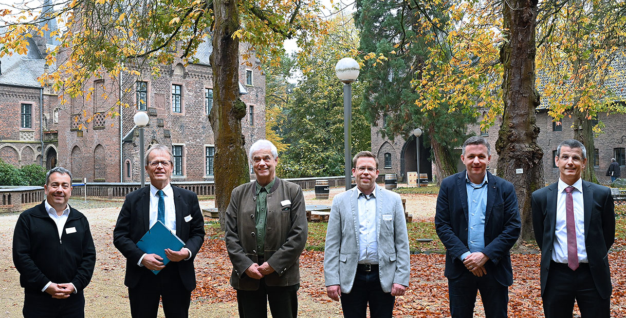 Eine Gruppe von sechs Männern steht im Freien vor einem historischen Gebäude, umgeben von herbstlichem Laub.