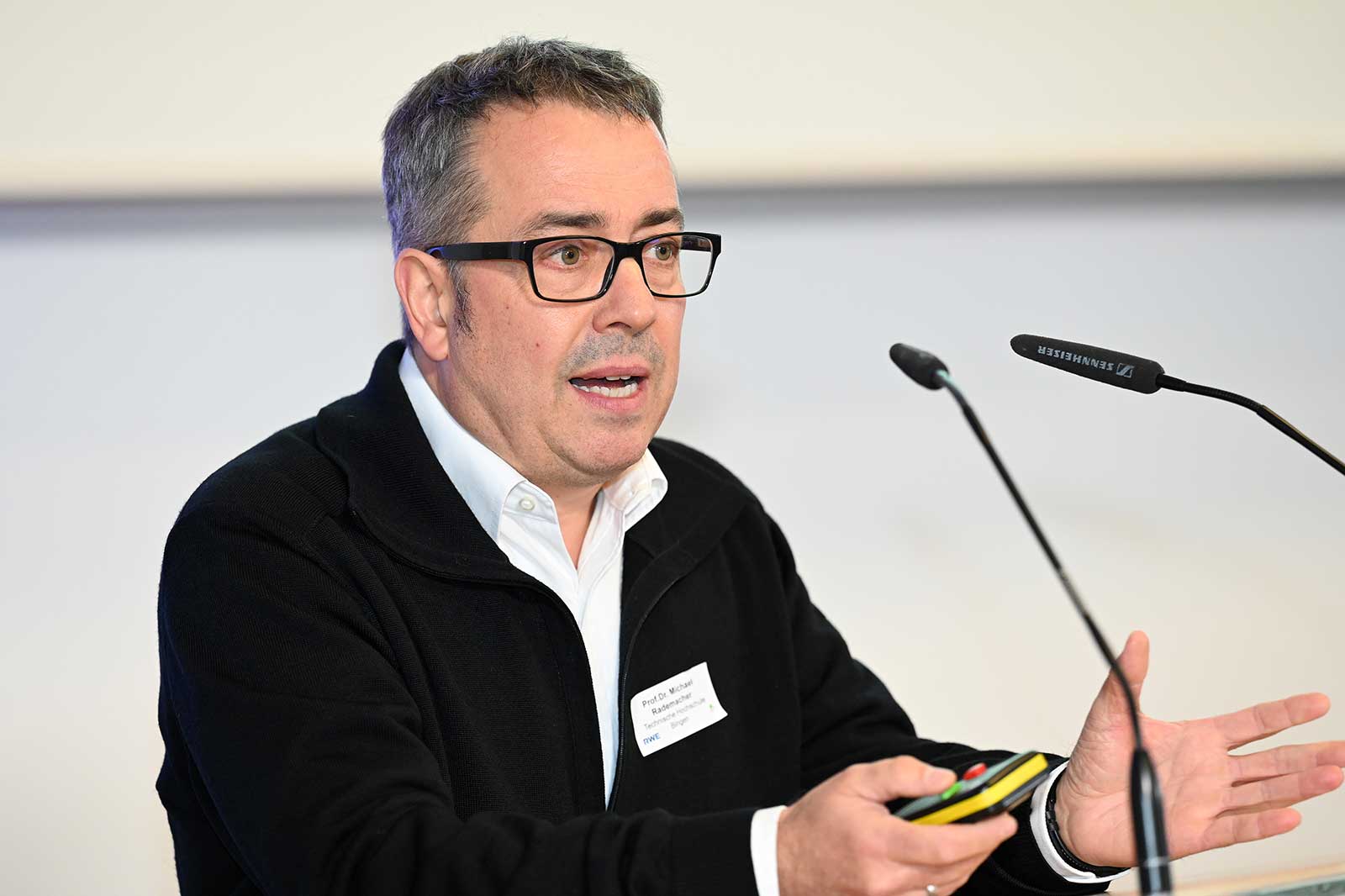 Ein Mann in einer schwarzen Jacke gestikuliert, während er eine Präsentationsfernbedienung bei einer Konferenz hält.