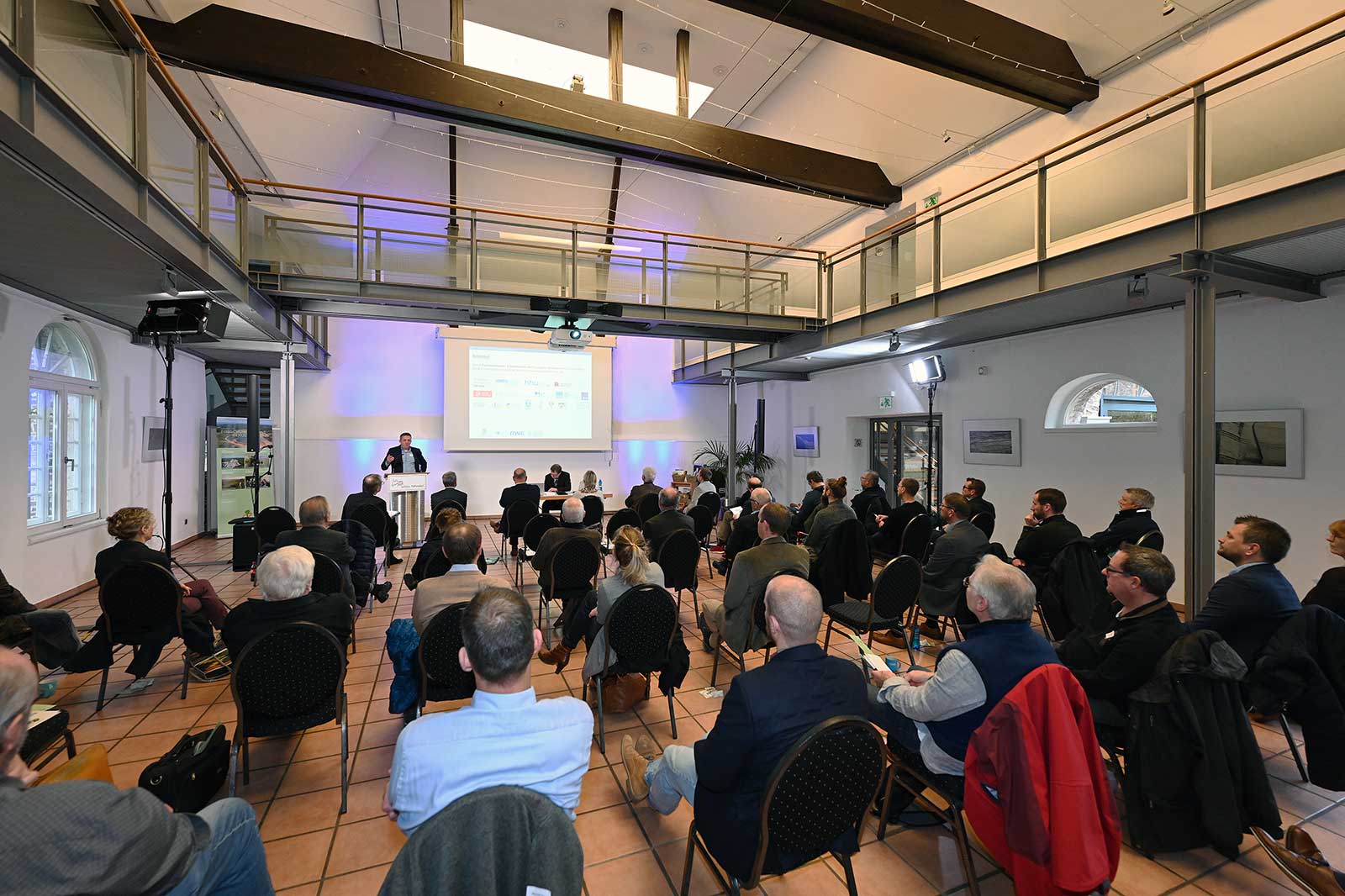 Ein Treffen in einem modernen Veranstaltungsort mit teilnehmenden Personen, die sitzen. Ein Redner präsentiert vor einem großen Bildschirm.