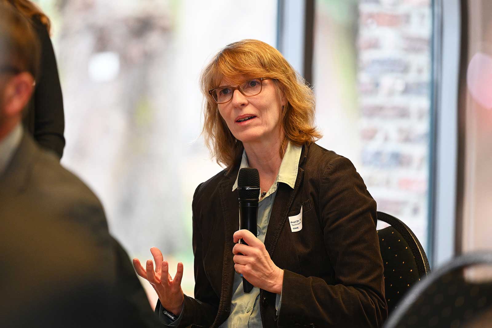 Eine Frau in einer dunklen Jacke hält ein Mikrofon und gestikuliert, während sie bei einer Veranstaltung in einer hellen Umgebung spricht.