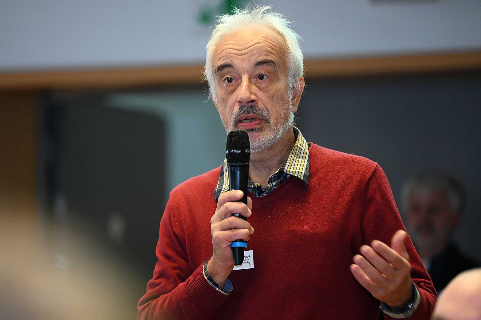 Ein älterer Mann in einem roten Pullover spricht in ein Mikrofon bei einer Veranstaltung und betont seine Punkte mit Gesten.