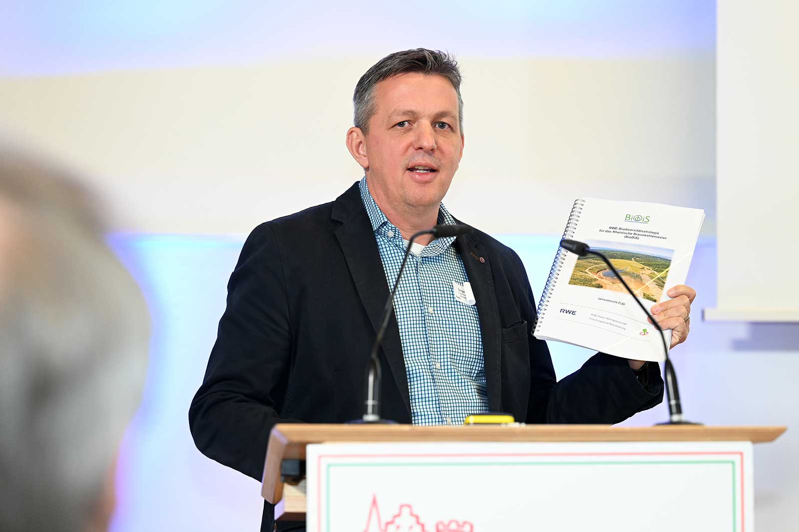 Ein Präsentator an einem Podium zeigt einen Bericht mit dem Titel 'RWE: Biodiversitätsstrategie für die Energiewende'.