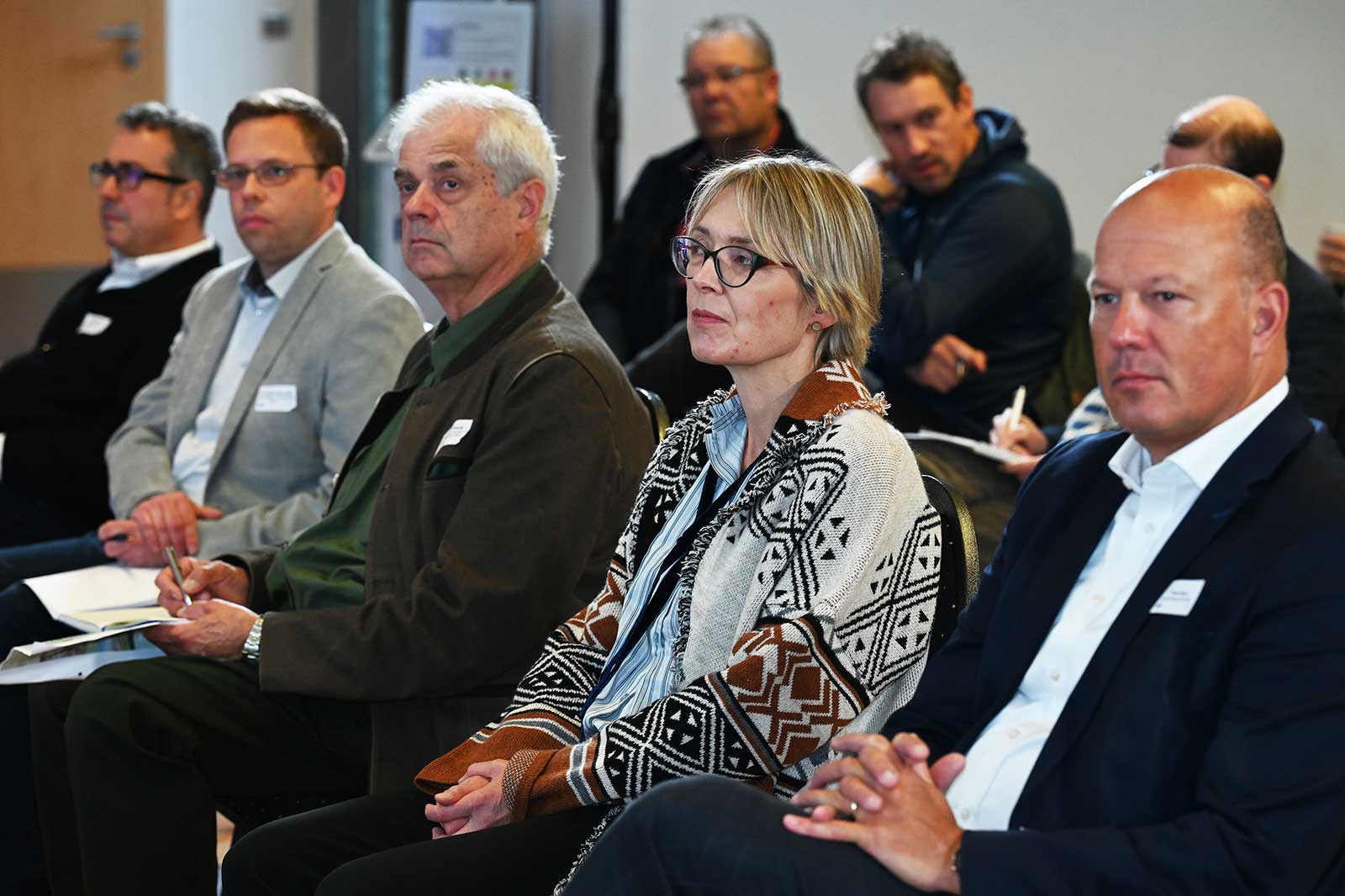 Eine Gruppe von Personen sitzt in einem Konferenzraum und ist an einer Präsentation oder Diskussion beteiligt, Notizen sind sichtbar.