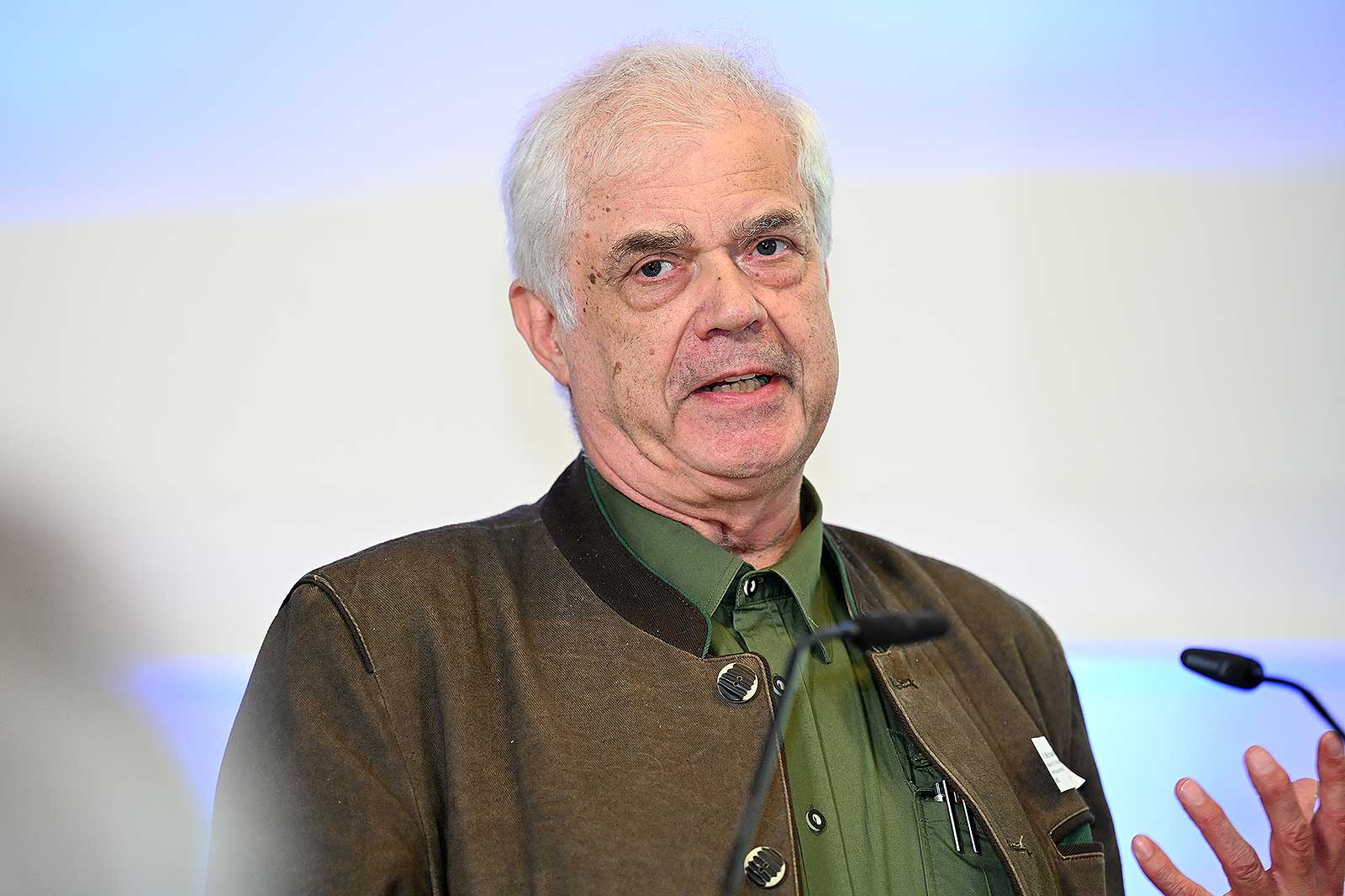 Eine Person in einer braunen Jacke spricht leidenschaftlich auf einer Konferenz und gestikuliert mit den Händen vor einem verschwommenen Hintergrund.