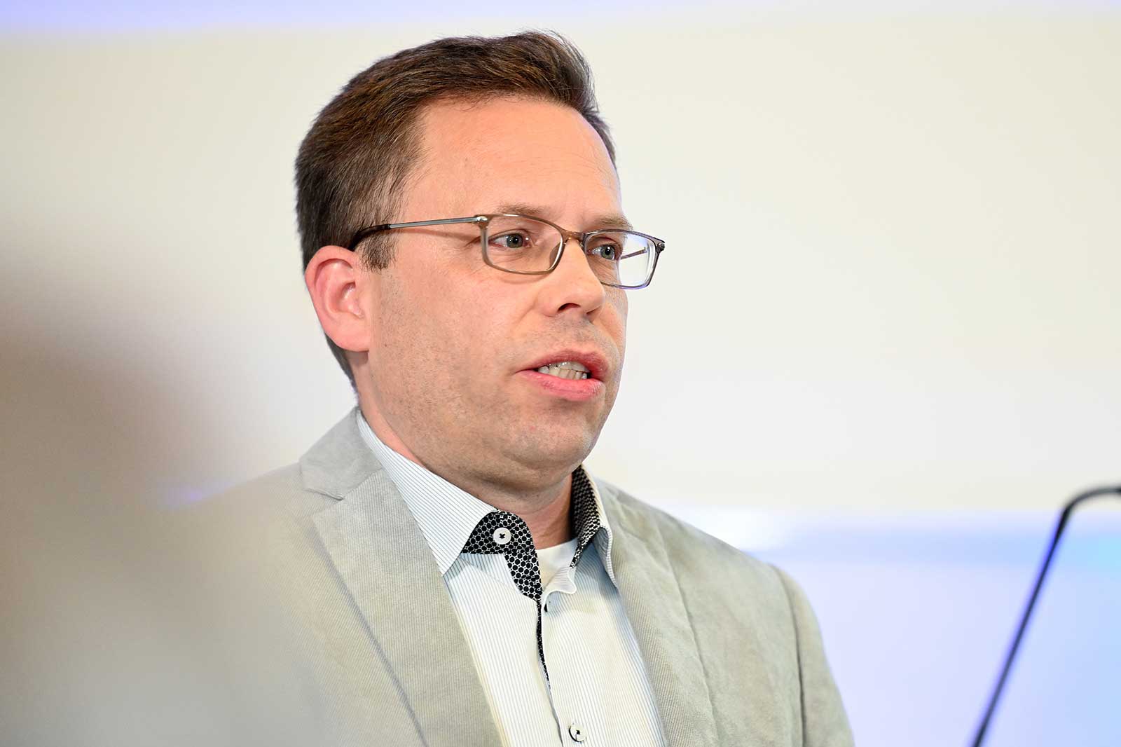 Ein Mann in einem grauen Blazer und einem gemusterten Hemd steht und blickt in eine unsichtbare Menge in einem professionellen Umfeld.