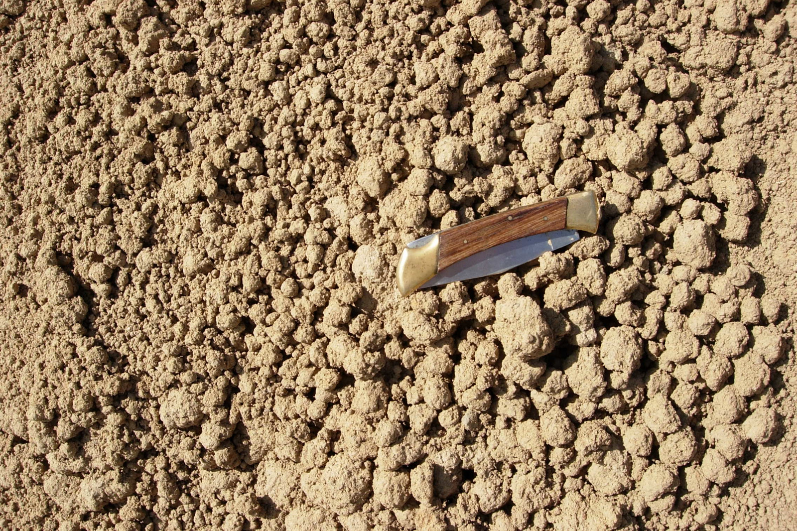 Ein Messer mit Holzgriff liegt auf einer strukturierten Oberfläche aus trockenem, rissigem Erdreich mit kleinen Klumpen und Erde.
