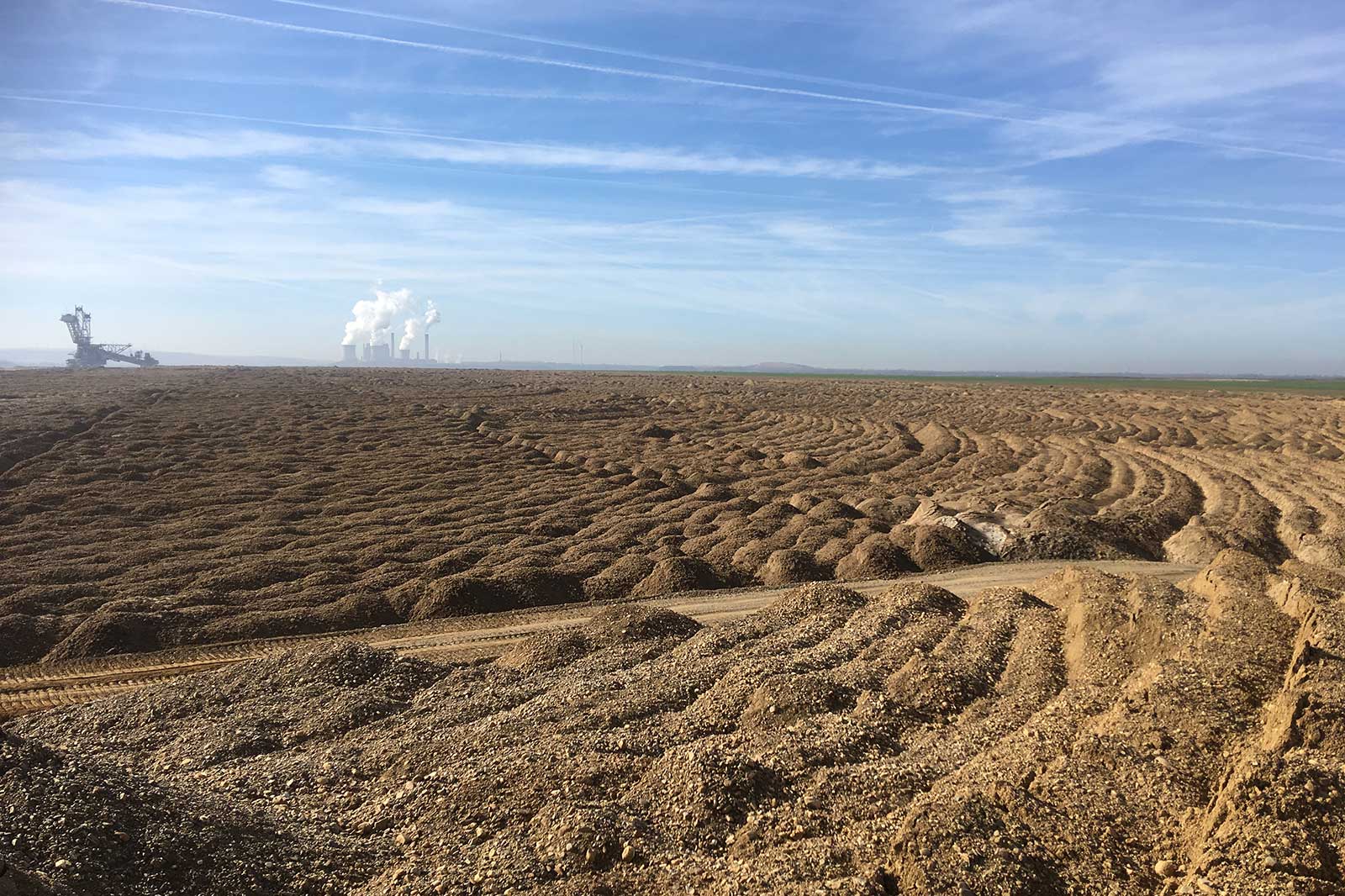 Eine weite, strukturierte Landschaft mit Erdhügeln, Maschinen in der Ferne und Rauch, der von Kraftwerken unter einem klaren Himmel aufsteigt.