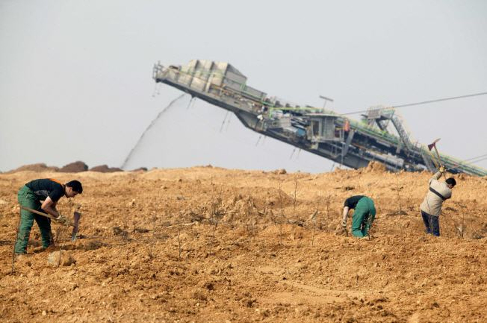 Arbeiter graben in einer kargen Landschaft mit einer großen Maschine im Hintergrund, die anscheinend Materialien ausgräbt.