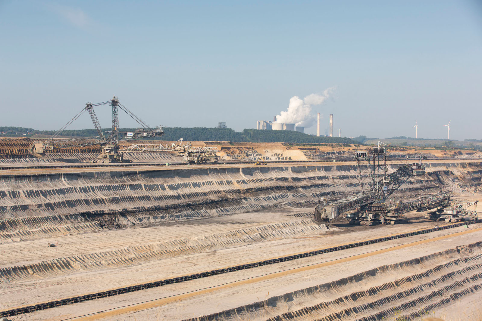Eine große Braunkohlenabbaustelle mit mehreren Baggern und einer rauchenden Industrieanlage im Hintergrund.
