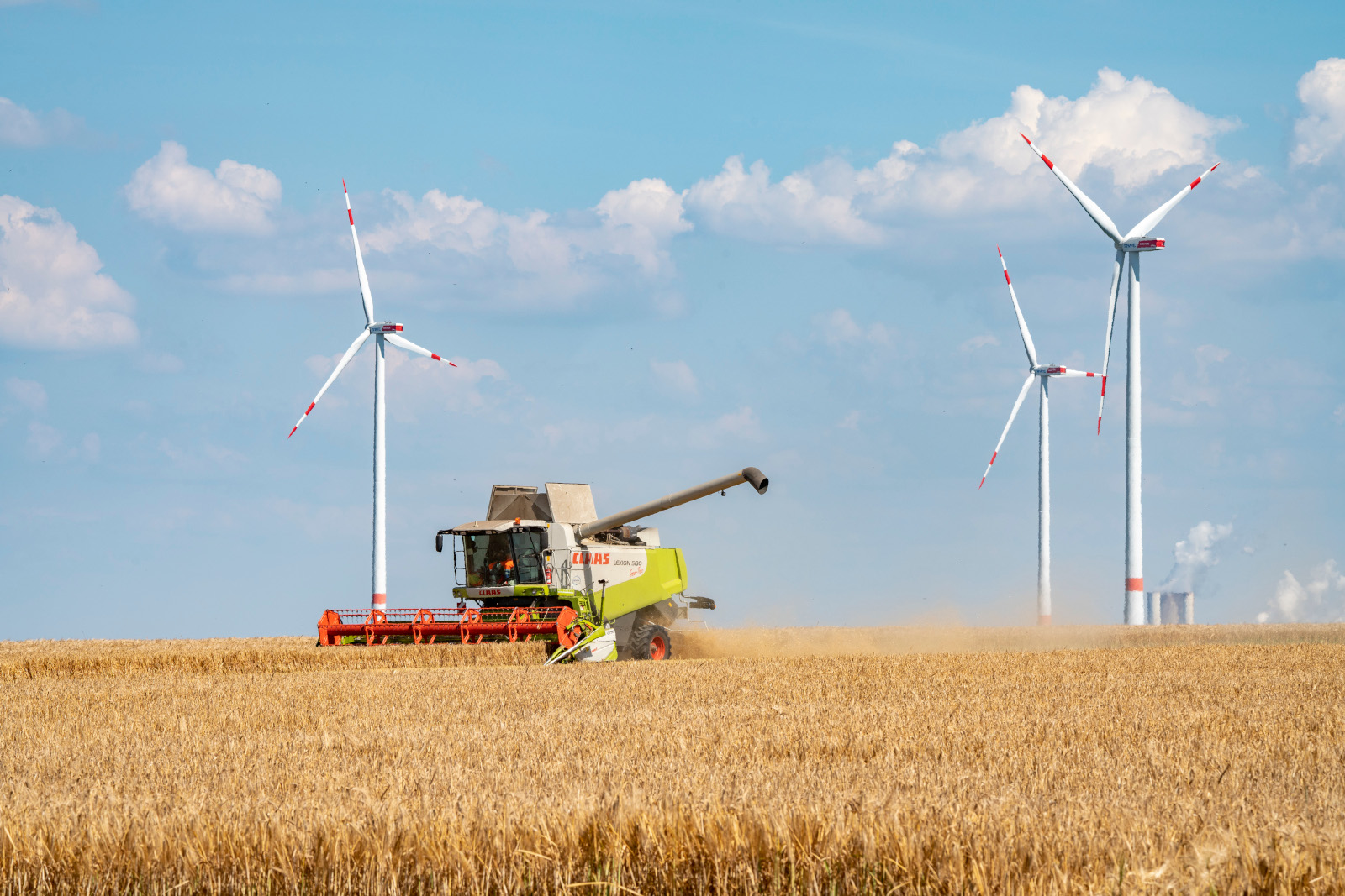 Ein Mähdrescher arbeitet in einem goldenen Feld, mit Windkraftanlagen im Hintergrund und einem blauen Himmel voller Wolken.
