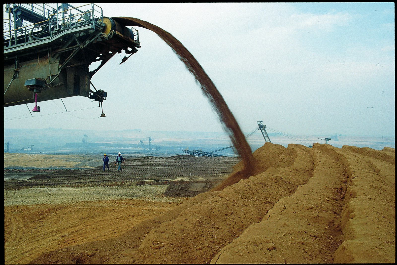 Eine große Maschine entlädt braunes Material auf einer Baustelle, während zwei Arbeiter im Vordergrund beobachten.
