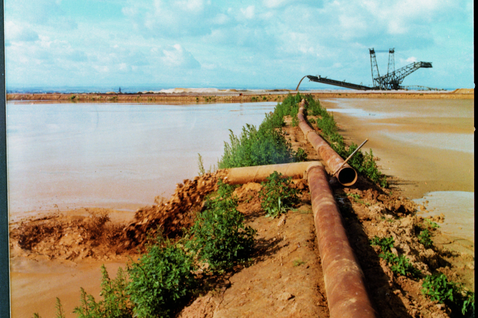 Eine Landschaft mit schlammigem Wasser, großen Rohren, grünen Sträuchern und einer Abbaumachine im Hintergrund unter einem bewölkten Himmel.