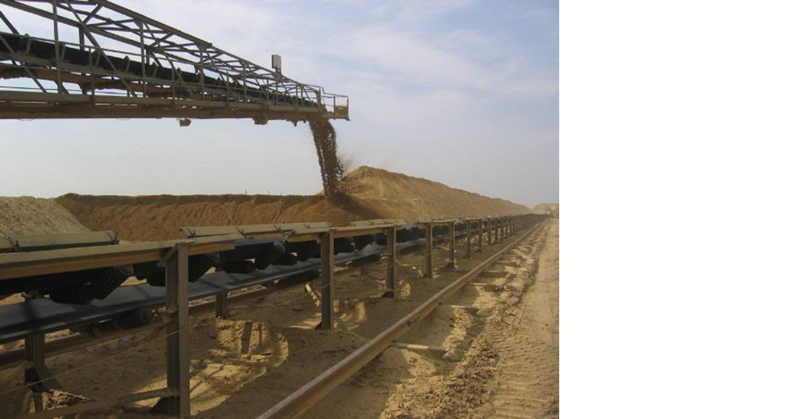 Ein Förderbandsystem transportiert Sand, mit einem Sandhaufen im Hintergrund unter einem klaren Himmel.