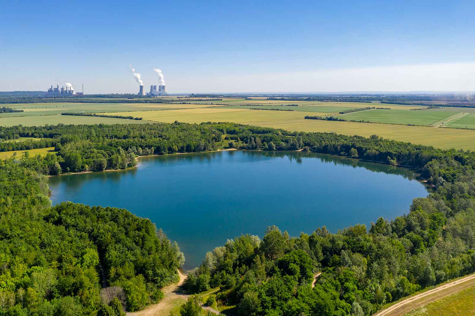 Luftaufnahme eines ruhigen Sees, umgeben von dichter Grünfläche, mit sichtbaren Industrieanlagen im Hintergrund unter einem klaren blauen Himmel.