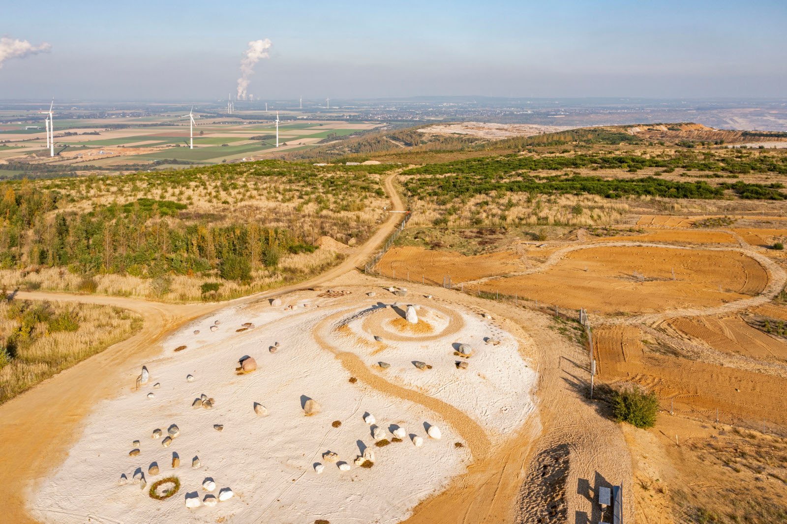 Eine Vogelperspektive auf eine Landschaft mit einer kreisförmigen Stein-Anordnung, Schotterwegen und Windrädern am Horizont.