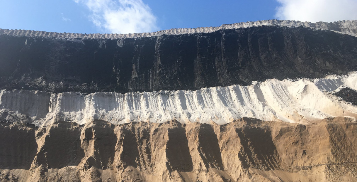 Eine geschichtete geologische Formation zeigt schwarze, weiße und braune Sedimente, die sich vor einem blauen Himmel abheben.