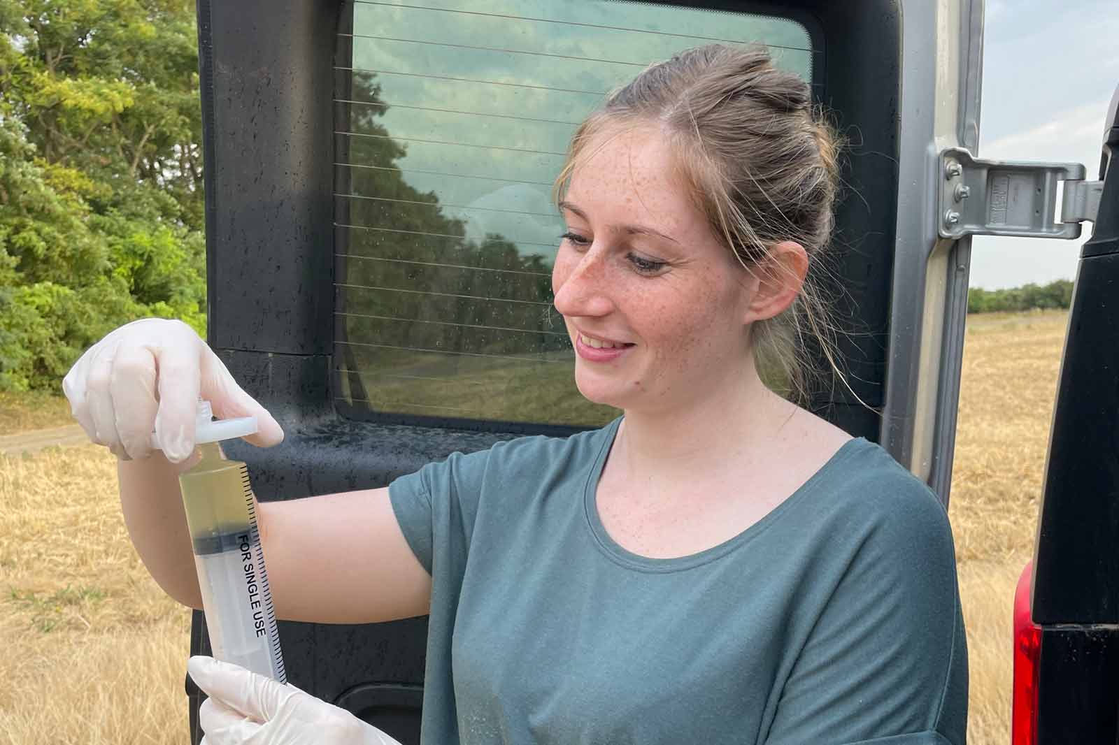 Eine Frau in einem grauen Shirt hält eine Spritze mit einer dunklen Substanz, trägt Handschuhe und steht im Freien neben Bäumen.