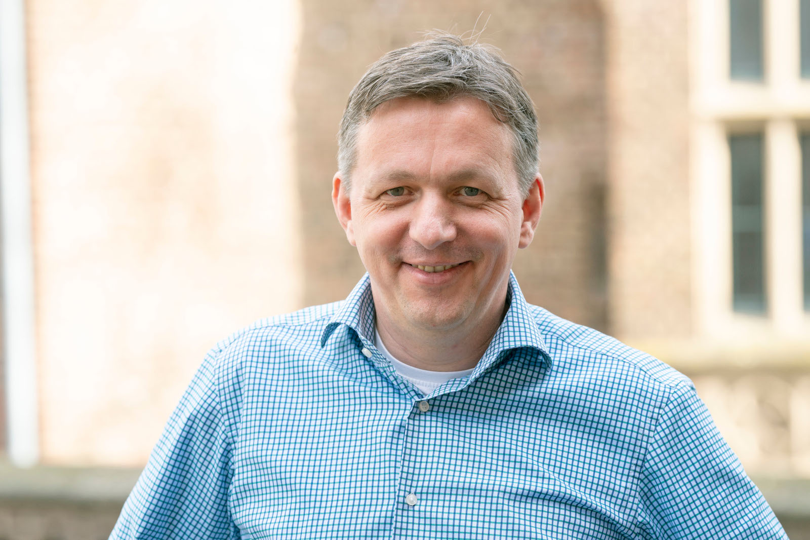 Ein Mann trägt ein kariertes Hemd und steht vor einem unscharfen Hintergrund mit Gebäuden.