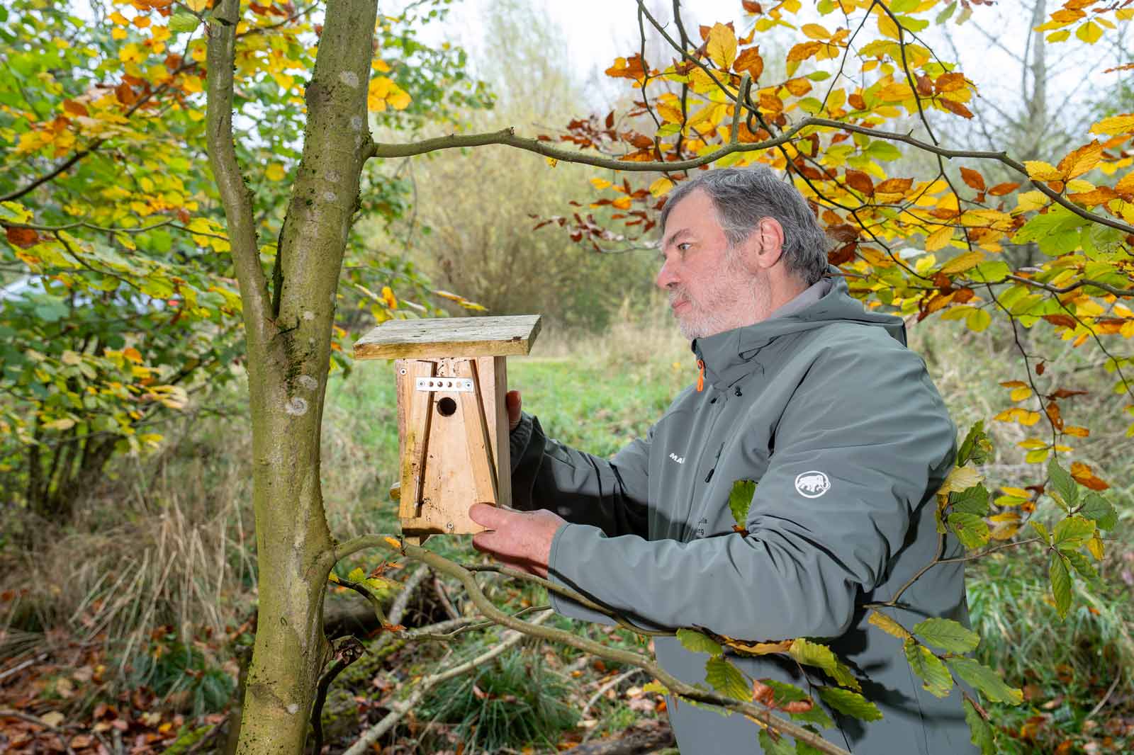 Ein Mann hält ein Vogelhaus aus Holz, das an einem Baum hängt, umgeben von buntem Herbstlaub.