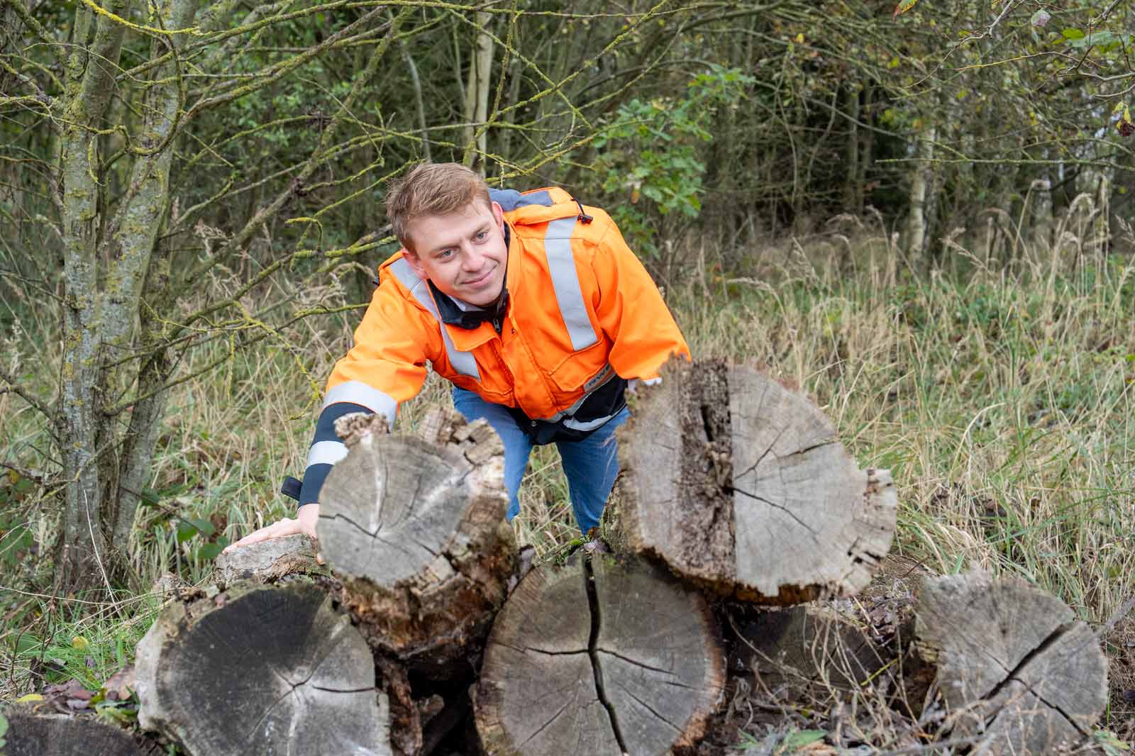 Ein Mann in orangefarbener Sicherheitsjacke beugt sich über einen Stapel Baumstämme in einem bewaldeten Bereich.
