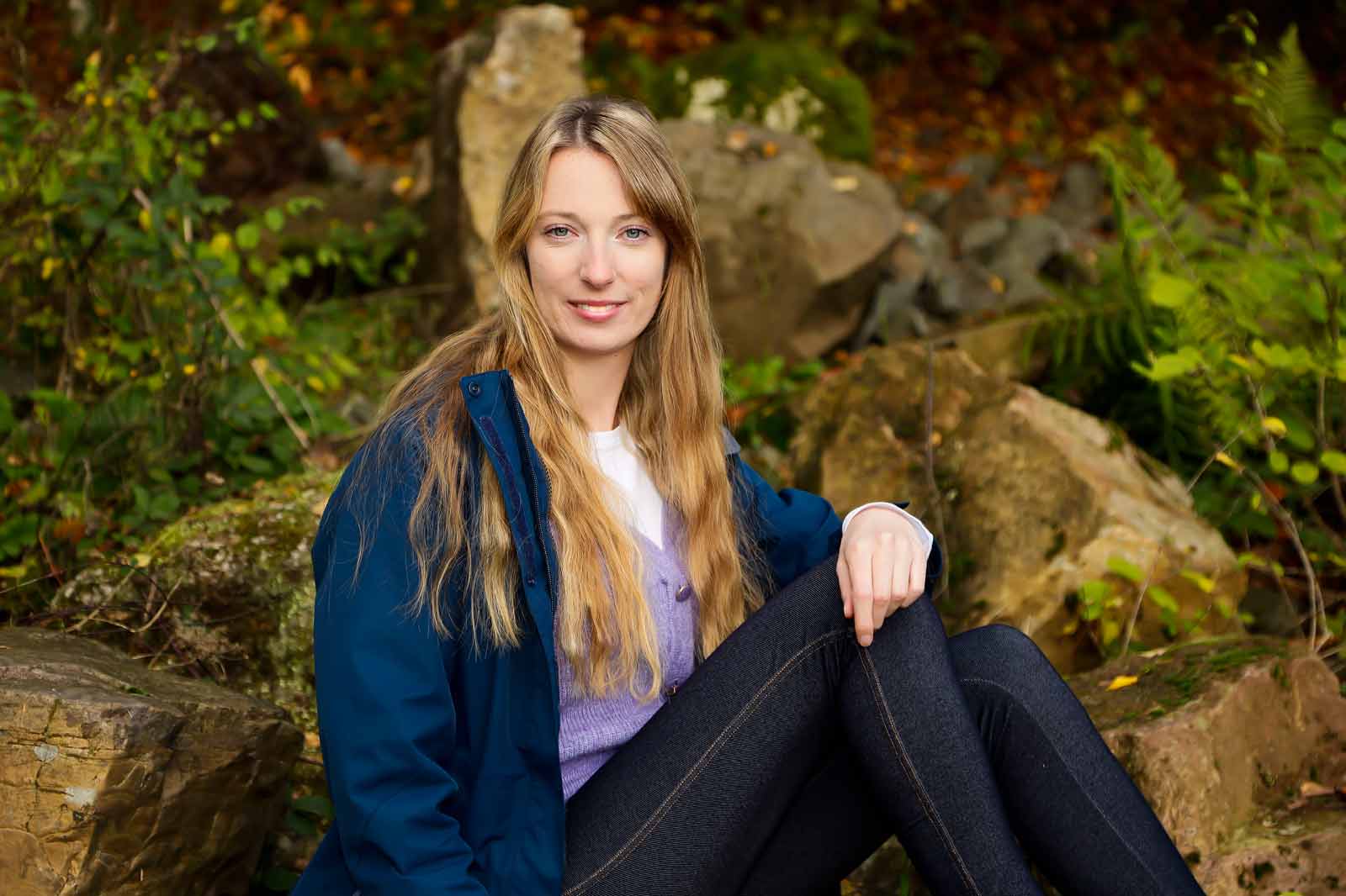 Eine Person mit langen, blonden Haaren sitzt auf Felsen in einer herbstlichen Umgebung, umgeben von Bäumen und Pflanzen.