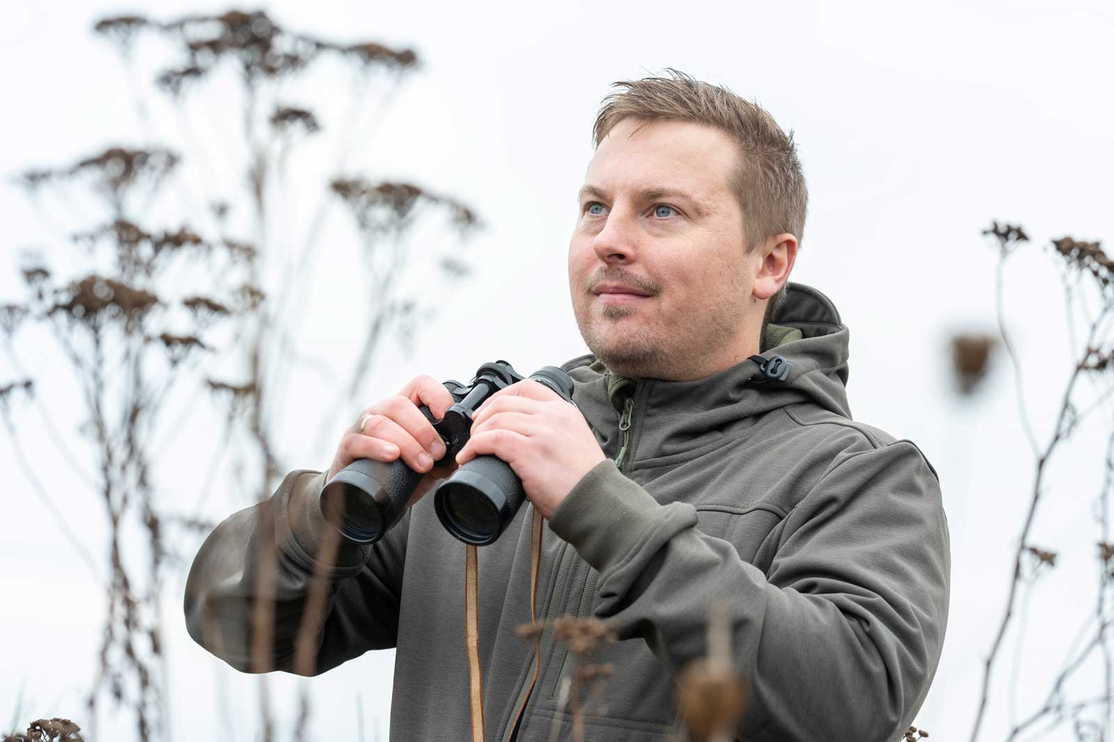 Eine Person hält ein Fernglas im Freien, umgeben von vertrockneten Blumen vor einem grauen Himmel.